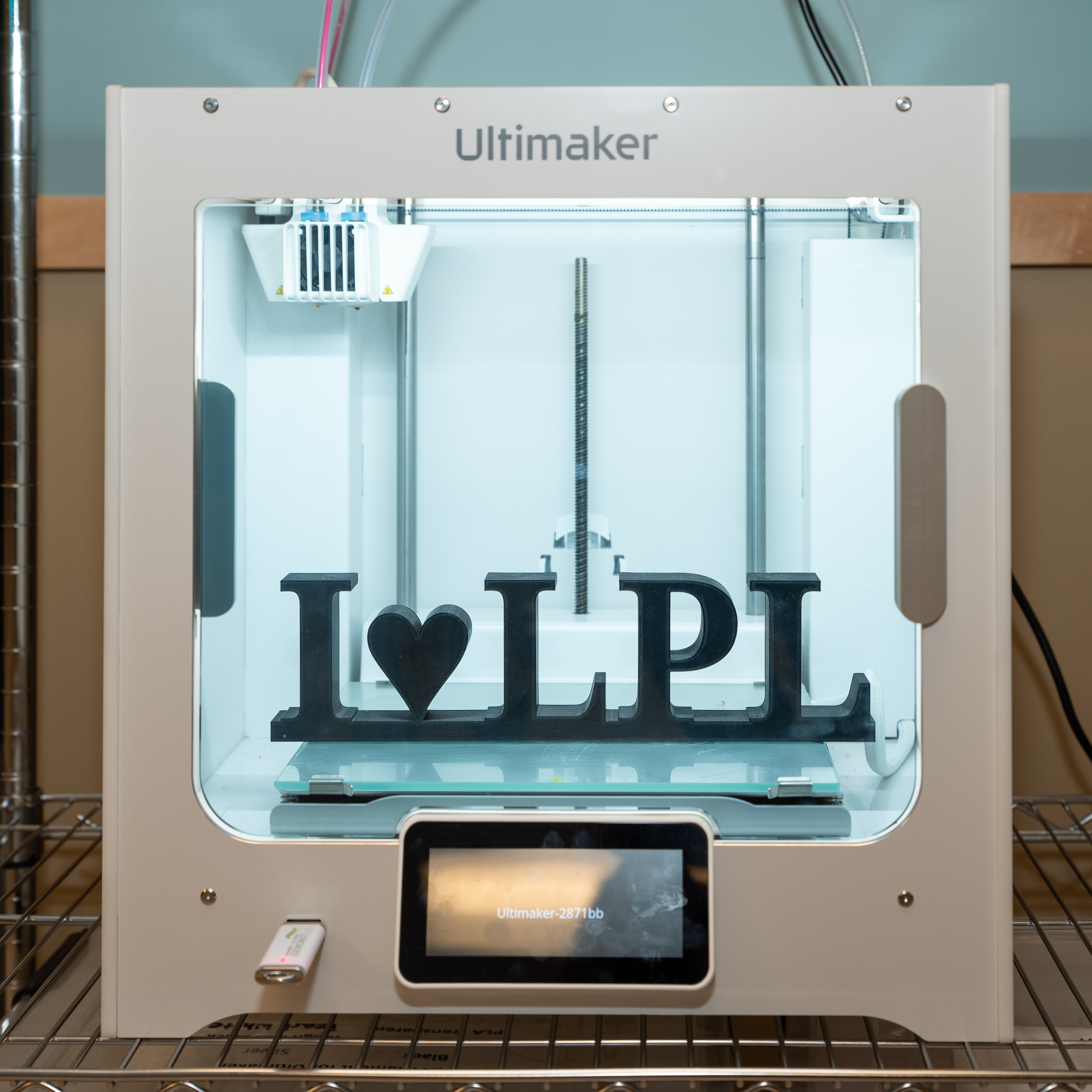 A white Ultimaker 3D printer inside the library’s maker space prints a black “I ♥ LPL” sign on the build plate, visible through the printer’s glass front.